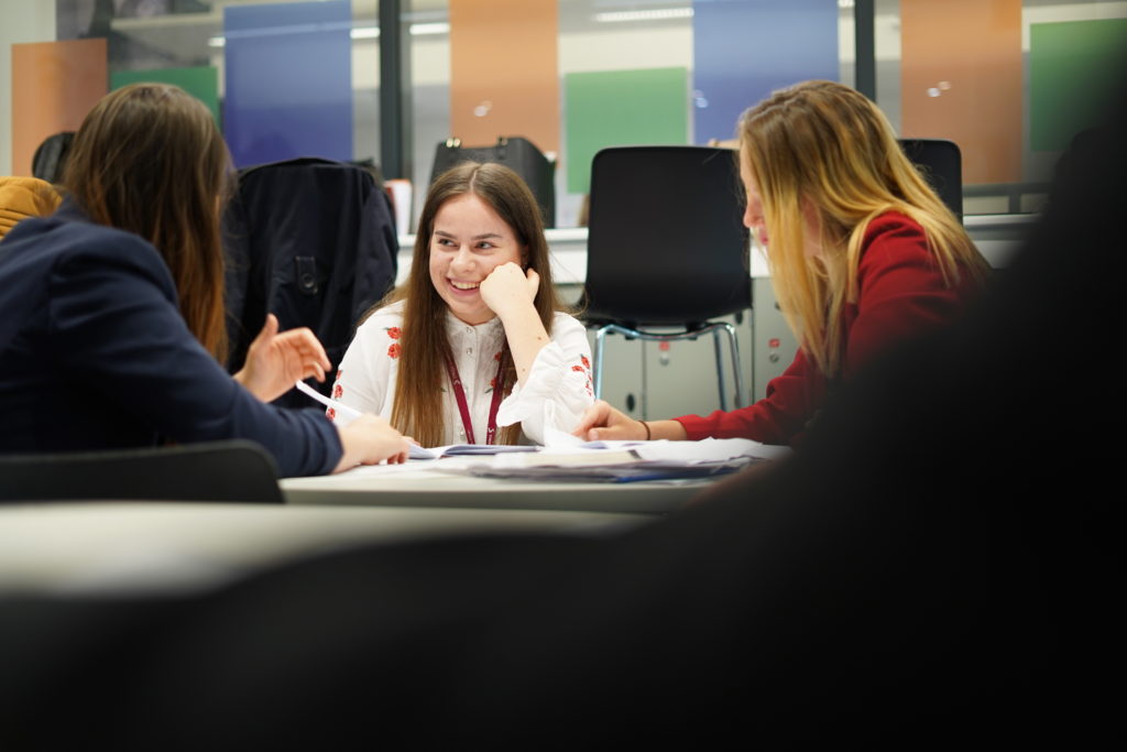 A small group of Post-16 students are seen sitting, working together at a desk and conversing with one another.