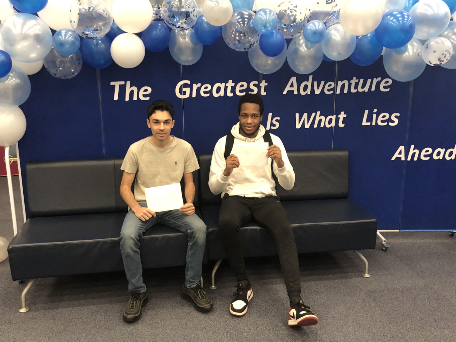 Two Post-16 students are pictured sitting next to each other on a bench, holding their A Level Results sheets in their hands and smiling for the camera.