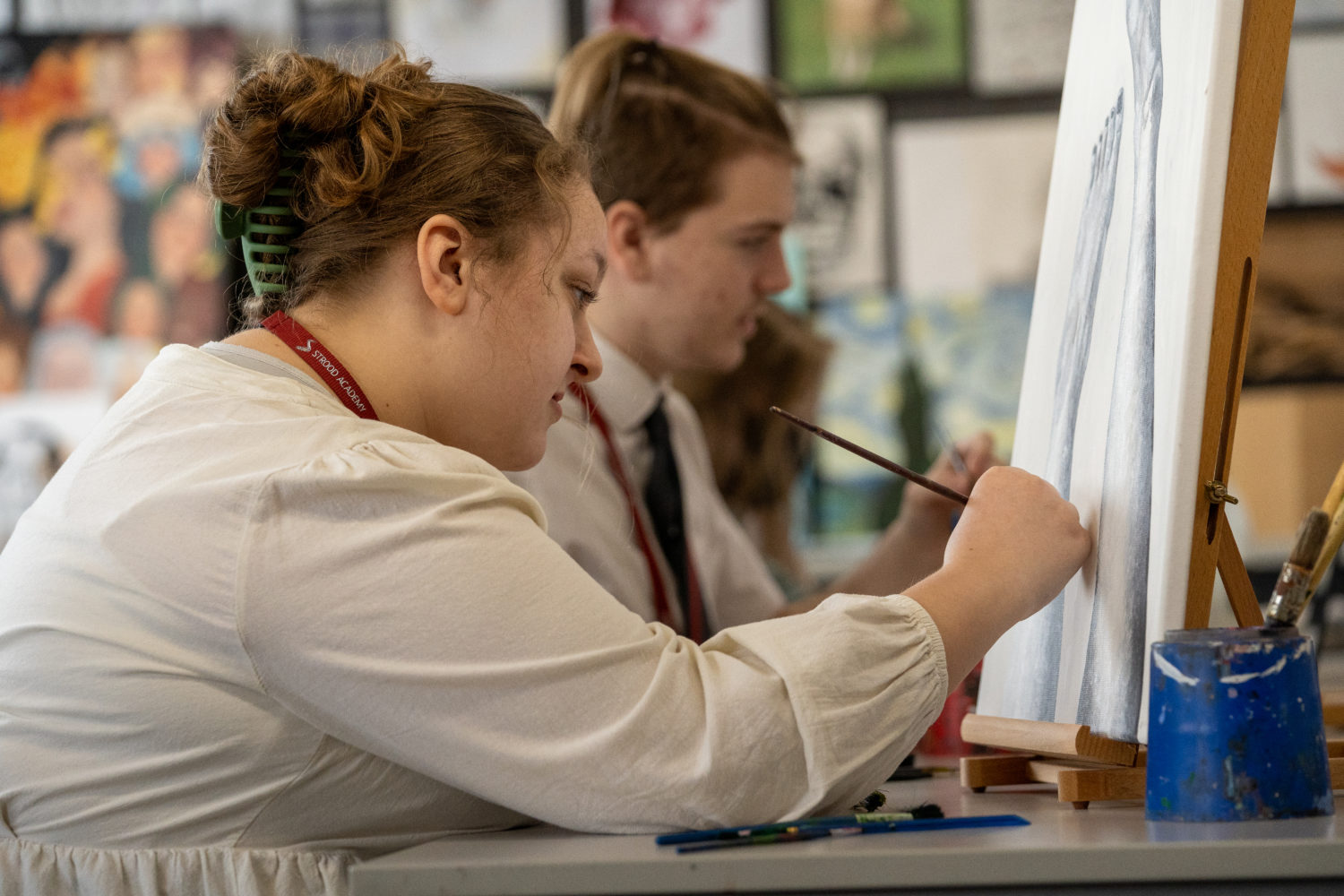 A female student painting on an easel with a male student in the background also painting
