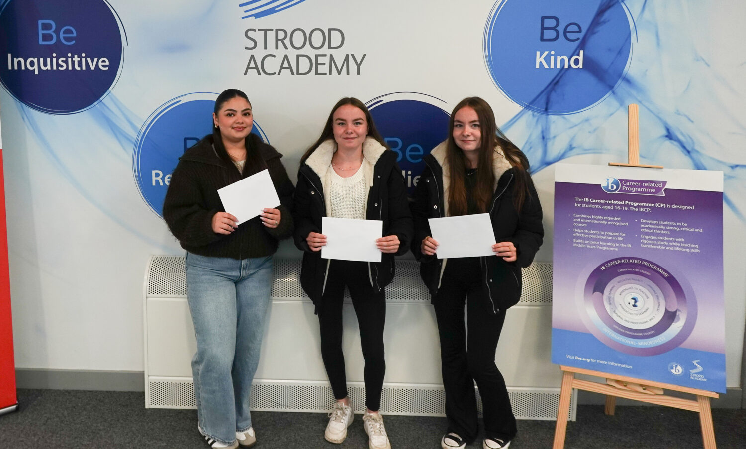 Three young women are standing side-by-side, each holding white papers, likely their results. They are in front of a wall with "STROOD ACADEMY" prominently displayed, along with positive attributes like "Be Inquisitive," "Be Kind," "Be Respectful," and "Be Resilient." There is a poster about "The IB Career-related Programme (CP)."