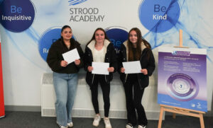 Three young women are standing side-by-side, each holding white papers, likely their results. They are in front of a wall with "STROOD ACADEMY" prominently displayed, along with positive attributes like "Be Inquisitive," "Be Kind," "Be Respectful," and "Be Resilient." There is a poster about "The IB Career-related Programme (CP)."