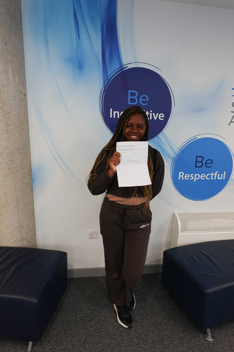A young woman is smiling and holding up a document (likely her results) in front of her. She is standing in the same Strood Academy setting as the previous photo, with the "Be Inquisitive" and "Be Respectful" circles visible behind her.